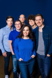 Los seis miembros del grupo sonríen a la cámara. Todos van vestidos de azul, con una mujer al frente. Los demás son hombres. 