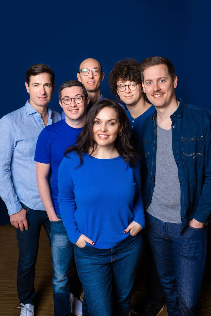 Los seis miembros del grupo sonríen a la cámara. Todos van vestidos de azul, con una mujer al frente. Los demás son hombres.